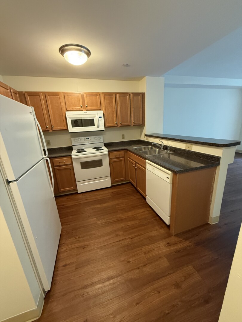 Kitchen with wood-tone cabinetry, dark countertops, white appliances including a refrigerator, electric range, microwave, and dishwasher, plus a breakfast bar with seating space.