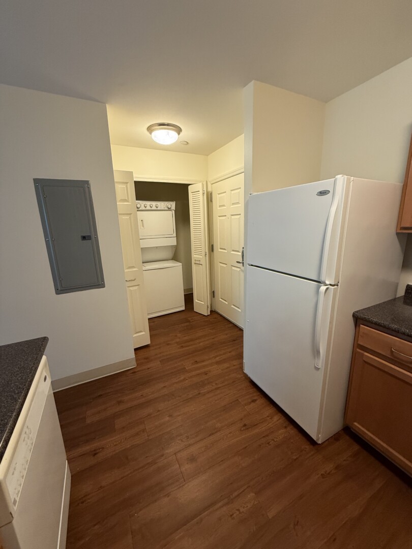 Kitchen view showing wood-tone cabinets, white refrigerator, dark countertops, and a stacked washer and dryer located in a nearby laundry closet.