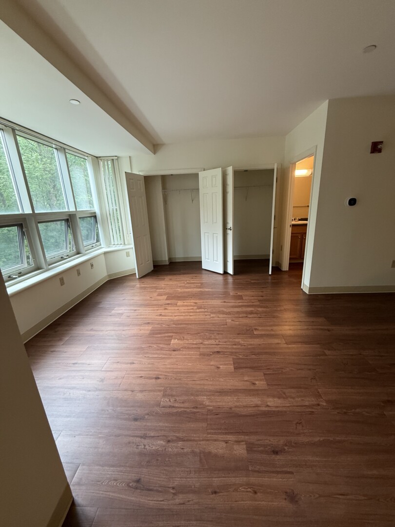 Bright room featuring large windows with wooded views, dark wood-look flooring, two closets, and a bathroom entrance. A breakfast bar separates the room from the kitchen area.
