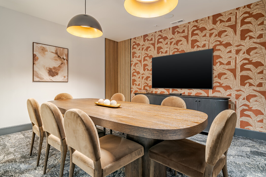 Conference room with patterned carpet flooring, light-colored walls, a white ceiling, a wood table, upholstered chairs, and a wall-mounted television.