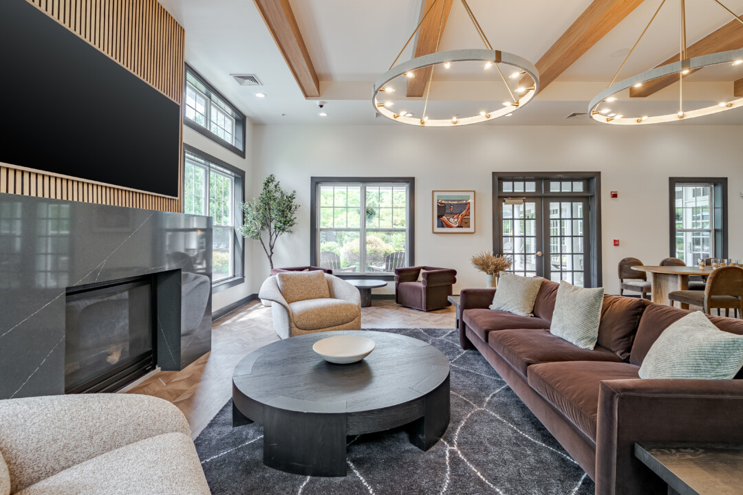 Community lounge with wood-look flooring, white walls, a white ceiling with wood beams, large windows, a dark fireplace surround, and upholstered seating.