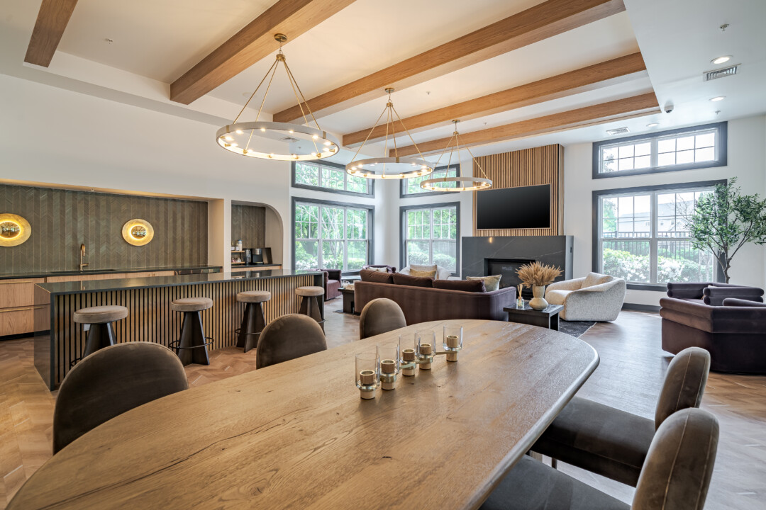 Community lounge with wood-look flooring, white walls, a white ceiling with wood beams, a dining table, bar seating, and large windows.