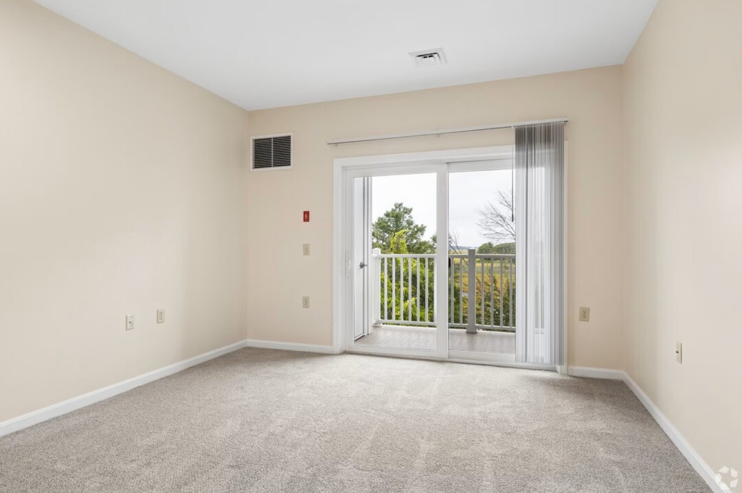 Empty living room with beige walls, light gray carpeting, and a sliding glass door leading to a private balcony. Vertical blinds partially cover the door, and the balcony overlooks green trees and open marshland. Natural light fills the space, highlighting the clean, neutral finishes and open layout.