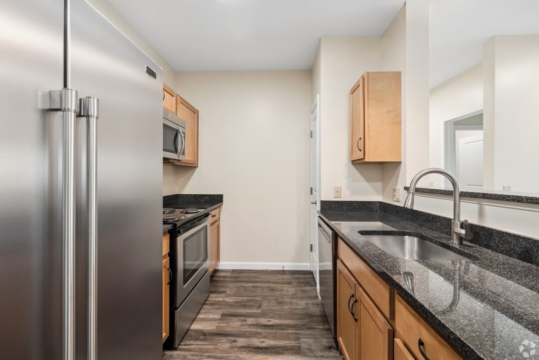 Galley-style kitchen with stainless steel appliances, including a refrigerator, electric range, microwave, and dishwasher. The kitchen features light wood cabinetry, dark granite countertops, a stainless steel sink with a high-arc faucet, and wood-look flooring. An open pass-through overlooks the adjacent living area, creating a bright and functional layout.