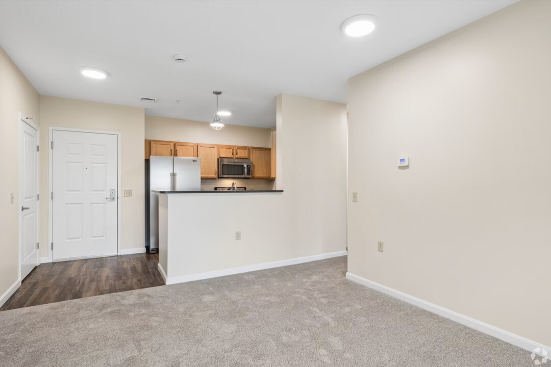 Open-concept living and dining area with light gray carpeting, beige walls, and recessed ceiling lights. The space opens to a kitchen featuring light wood cabinetry, stainless steel appliances, dark countertops, and a breakfast bar. A white entry door and wood-look flooring define the transition between the living area and kitchen, creating a bright and functional layout.