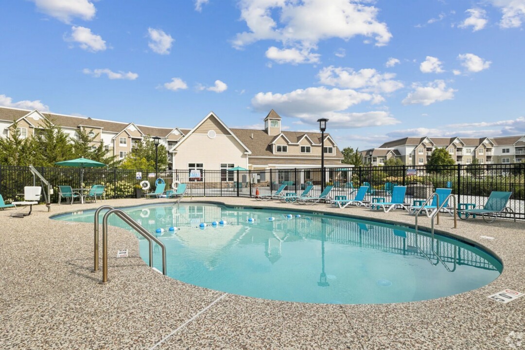 Outdoor swimming pool at an apartment community, surrounded by a fenced pool deck with turquoise lounge chairs, tables, and umbrellas. Multi-story residential buildings and a clubhouse-style amenity building are visible in the background. The pool area is well-maintained and set beneath a bright blue sky with scattered white clouds.
