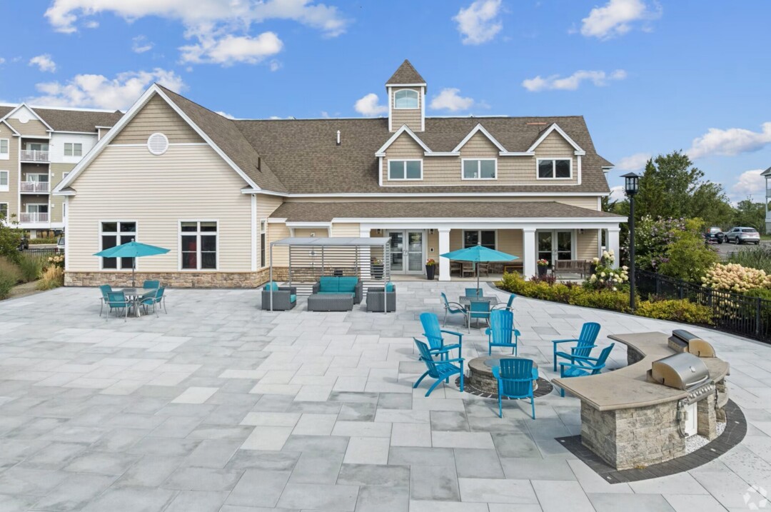 Apartment community clubhouse with a large outdoor patio featuring a circular fire pit, blue Adirondack chairs, dining tables with umbrellas, cushioned lounge seating, and a built-in grilling station. The clubhouse has beige siding, white trim, and a covered porch, with landscaped gardens surrounding the patio. Multi-story apartment buildings and a bright blue sky with scattered clouds are visible in the background.