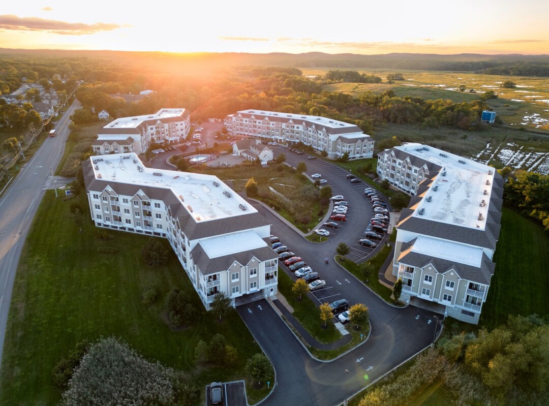 Aerial view of a large apartment community at sunset, featuring multiple four-story residential buildings arranged around landscaped grounds, parking areas, and a central clubhouse and pool. The property is bordered by expansive wetlands and wooded areas, with golden sunlight illuminating the buildings and surrounding landscape. A roadway runs alongside the community, and the horizon is framed by rolling hills beneath a warm evening sky.