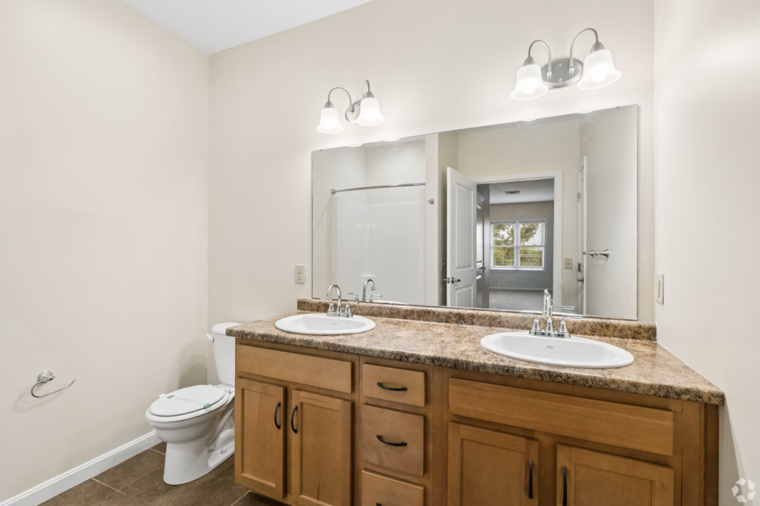 Spacious bathroom with a double-sink vanity featuring wood cabinetry, a granite-look countertop, and a large wall mirror. Two three-light vanity fixtures provide bright lighting above the sinks. The bathroom includes a toilet, tiled flooring, and a shower area reflected in the mirror. An open door leads to an adjacent bedroom with a window overlooking greenery.