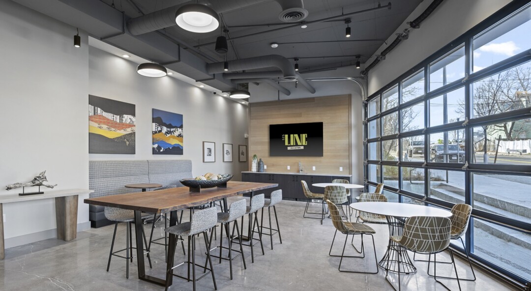Community room with white walls, a wood accent wall with a wall-mounted TV, dark-colored cabinets, seating, tables, and large windows.