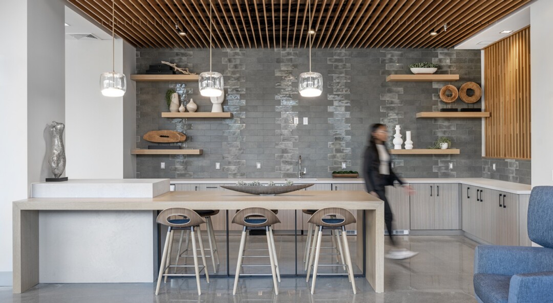 Community kitchen with a large island and bar stools, gray cabinets with a white countertop, a mix of gray tile and white walls, and a wood accent wall.