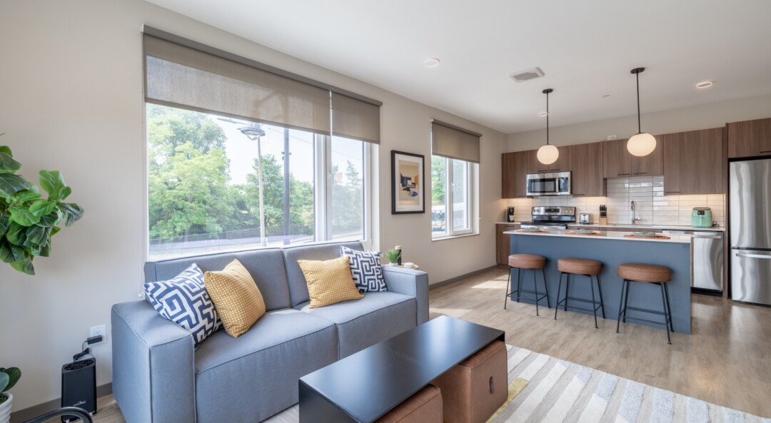 Open living room and kitchen with large windows, a gray sofa, and a breakfast bar with stools.