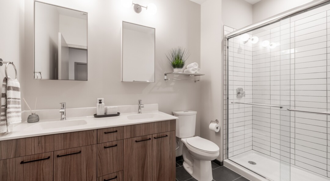 Bathroom with white walls, a double-sink wood vanity with mirrors and a white countertop, and a glass-enclosed shower with white tile next to a toilet.