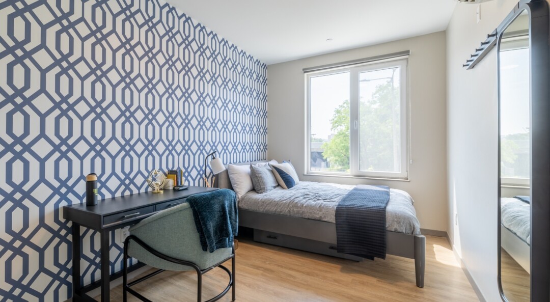 A bright bedroom with a large sliding glass window, white walls throughout, and one accent wall featuring printed designs.