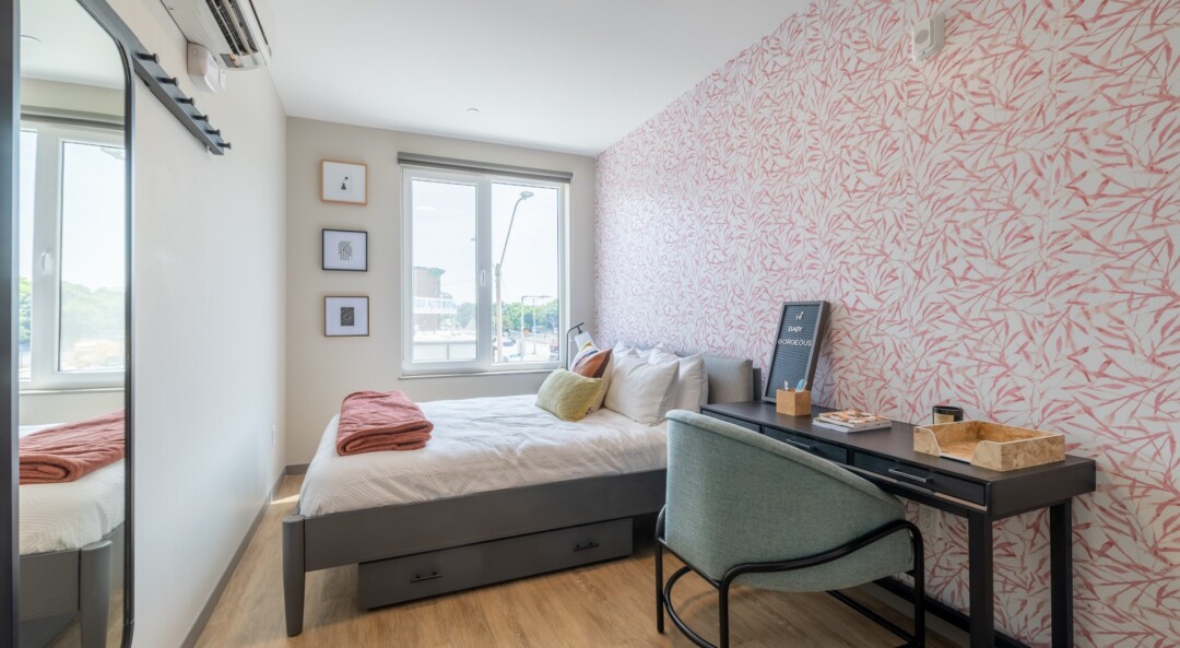 A bright bedroom with a large sliding glass window letting in natural light, white walls throughout, and one accent wall featuring printed designs that add visual interest.