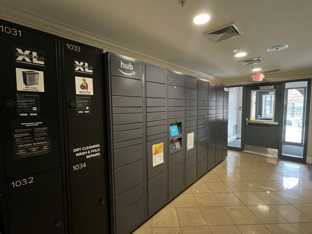 Indoor package drop off locker area with dark gray and black lockers; white ceiling with recessed lighting and vents, and glossy tan ceramic tile flooring near a glass entry door.