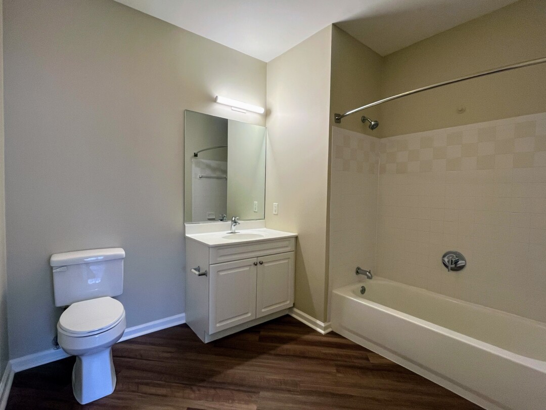 Bathroom with white toilet, white single-sink vanity, and light-colored countertop; beige walls, white ceiling, wood-style dark flooring, and a bathtub with off-white tiled surround and chrome fixtures.