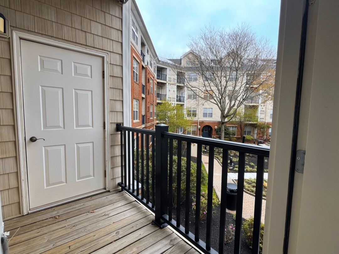 Outdoor balcony with natural wood plank flooring, black metal railing, a white door, and view of landscaped courtyard and nearby buildings.