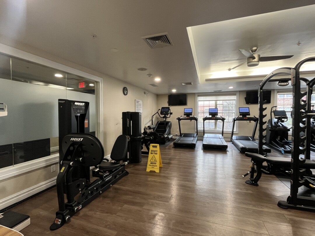 Spacious gym with wood-style flooring, neutral beige walls, and a white tray ceiling with recessed lighting and ceiling fans; features treadmills by windows, weight machines, and a mirrored wall.