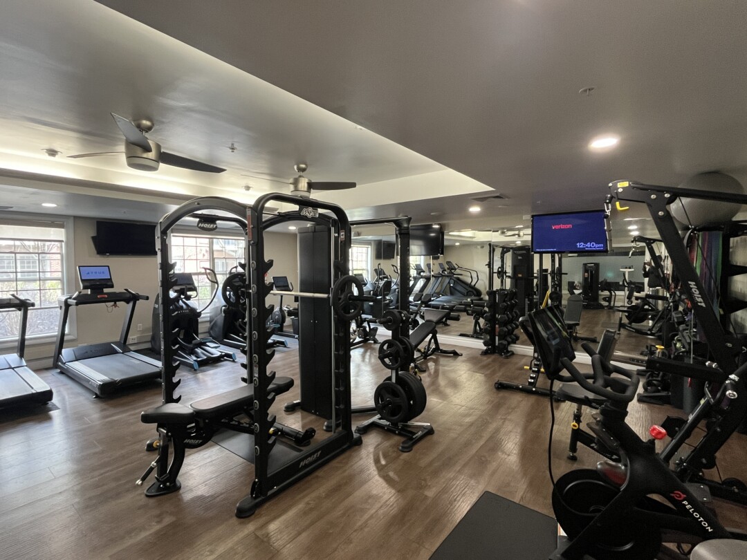Fitness center with wood-style flooring, light beige walls, and a white ceiling with recessed lighting and ceiling fans; includes black strength equipment, treadmills, and wall-mounted TVs with large mirrors along one wall.