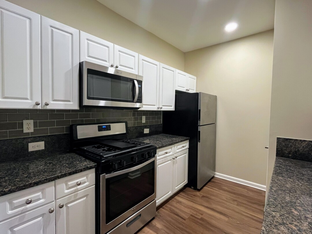 kitchen with white cabinets, dark speckled countertops, and gray tile backsplash; features stainless steel appliances, beige walls, white ceiling with recessed lighting, and wood-style flooring.