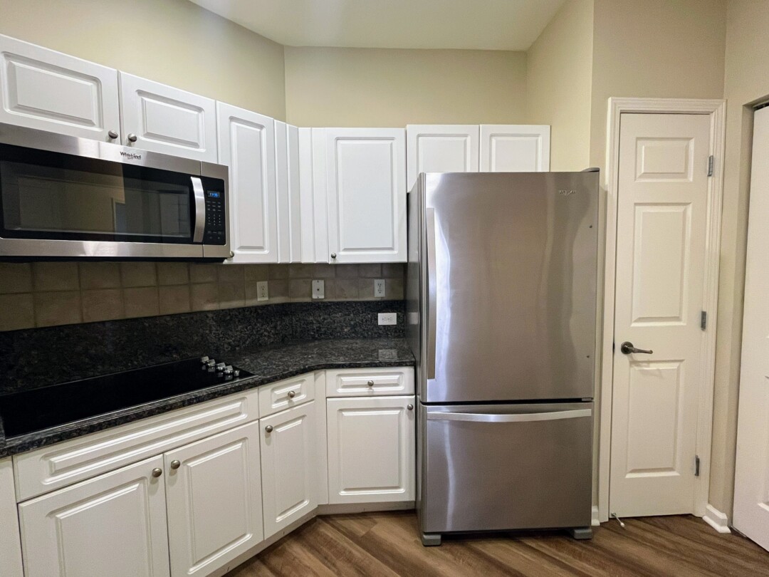 Kitchen with white cabinets, dark gray and black speckled countertops, beige tiled backsplash, stainless steel refrigerator and microwave, beige walls, and wood-style flooring.