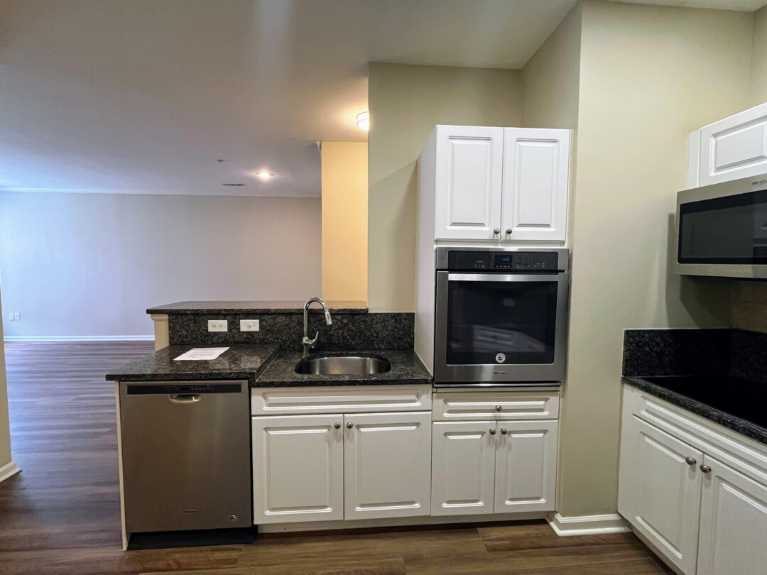 Kitchen with white cabinets, dark granite-style countertops, stainless steel sink and dishwasher, beige walls, white ceiling with recessed lights, and wood-style flooring open to living area.