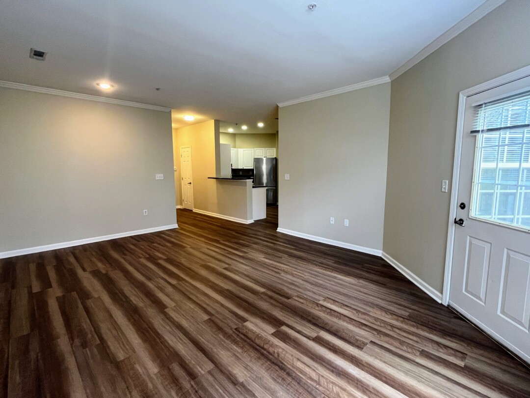 Open living area with dark wood-style flooring, light beige walls, white ceiling with recessed lighting, and view toward kitchen and hallway.