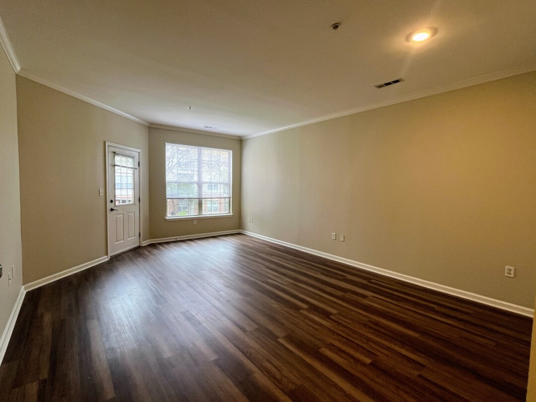 Large living room with dark wood-style flooring, light neutral walls, white ceiling, and window with white blinds next to a door leading outside.