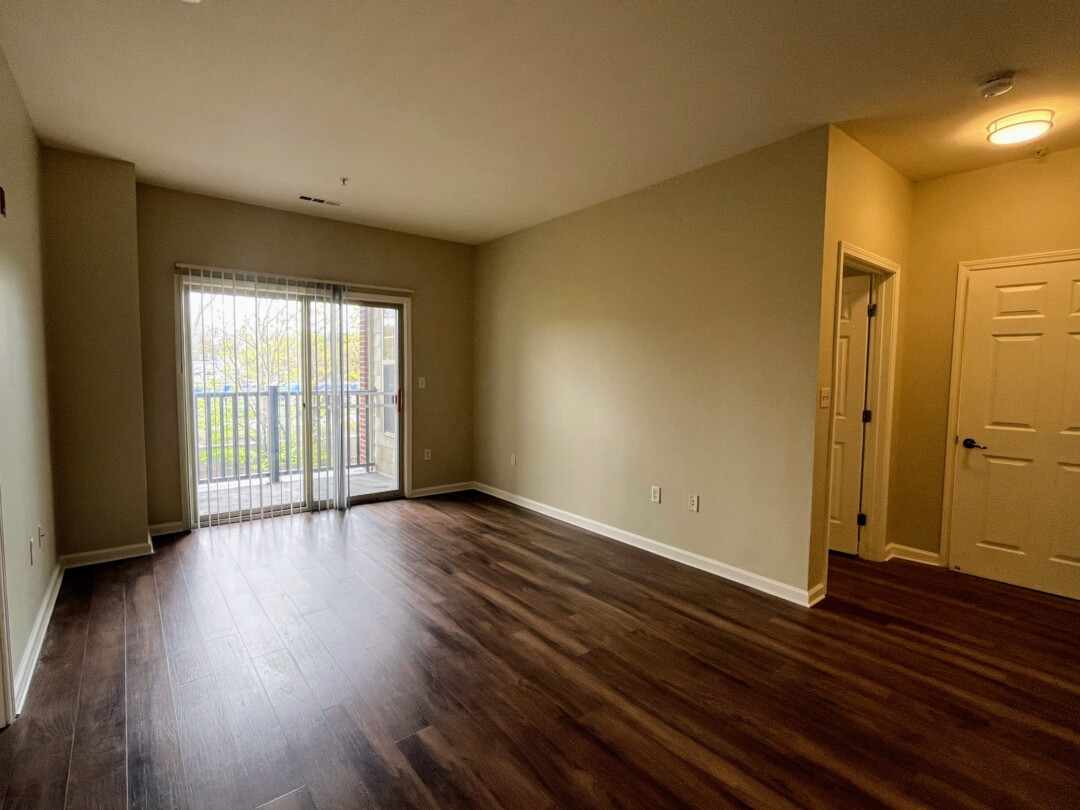 Open living room with beige walls, white ceiling, and dark wood-style flooring; includes a sliding glass door with vertical blinds leading to a private balcony and natural light.