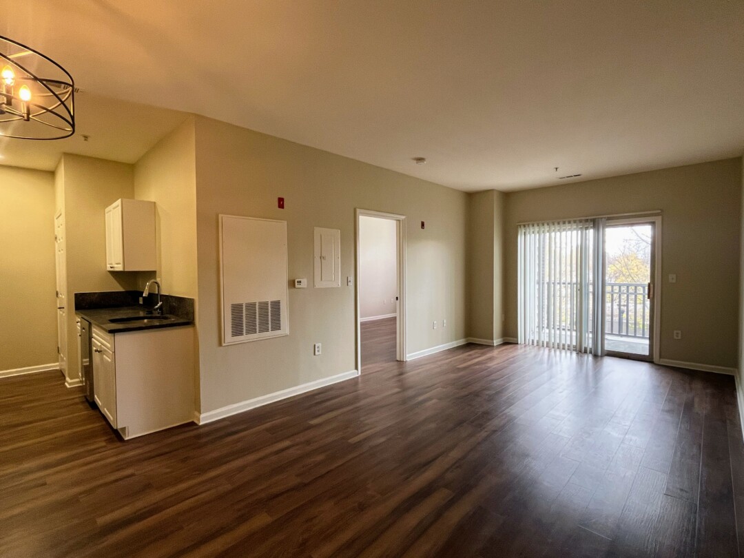 Spacious open-concept living area with beige walls, white ceiling, and dark wood-style flooring; includes a partial view of the kitchen, entry door, and sliding glass door to balcony with vertical blinds.