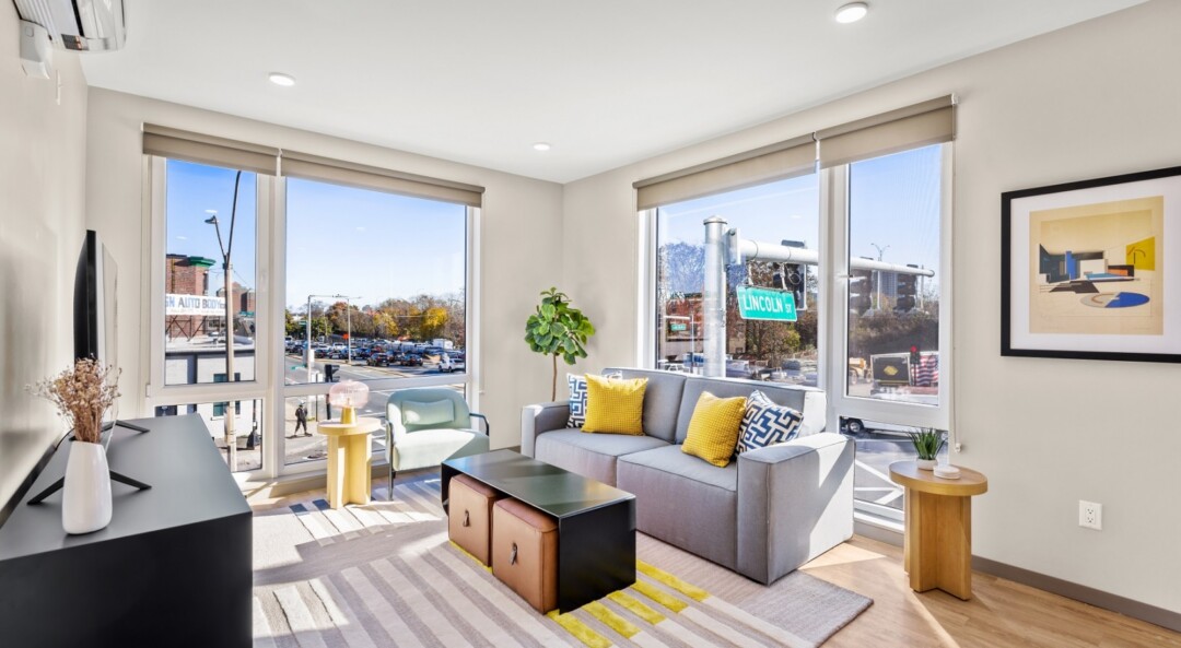 Fully furnished living room with white walls, large windows, a gray sofa, and natural light.