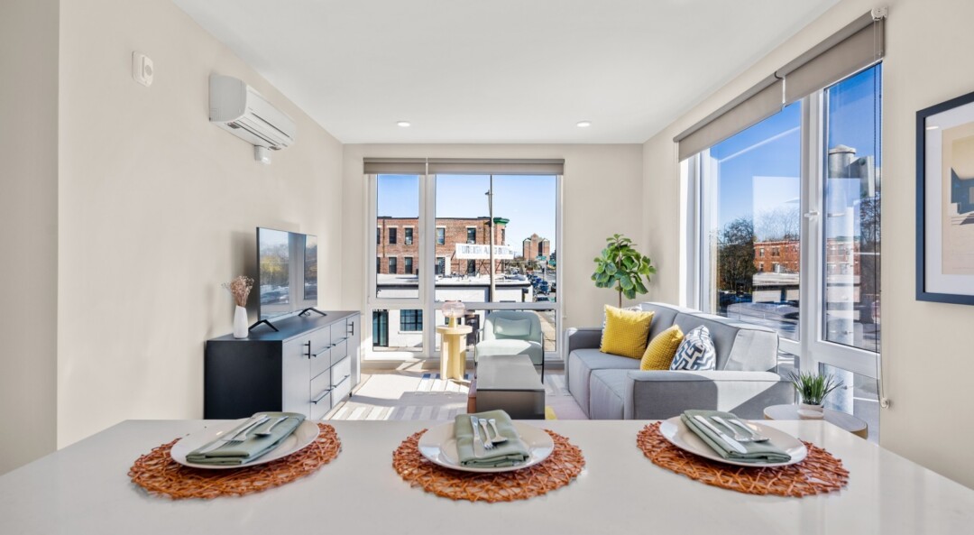 Apartment kitchen and living area with white walls and ceiling, large windows, and kitchen island with countertop seating.