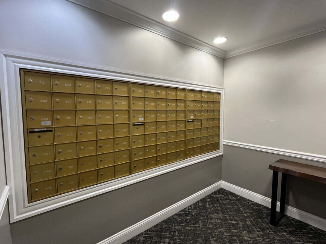 Mailroom with a wall of gold-colored metal mailboxes set into a white-trim frame; two-tone walls (light gray upper and darker gray lower), white ceiling with recessed lights, and dark patterned carpet flooring.