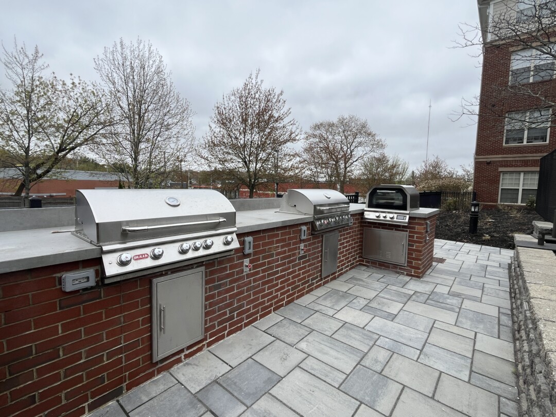 Outdoor grilling station with stainless steel grills set into red brick bases and gray stone countertops; surrounding gray stone paver flooring and landscaped area under an overcast sky.