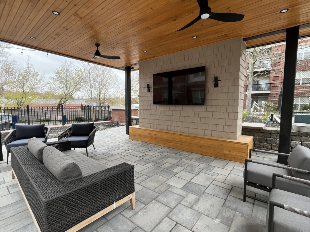 Covered patio lounge with gray stone paver flooring, wood-paneled ceiling with recessed lighting and black ceiling fans, and a central wall with mounted TV; furnished with cushioned seating in gray and black tones.