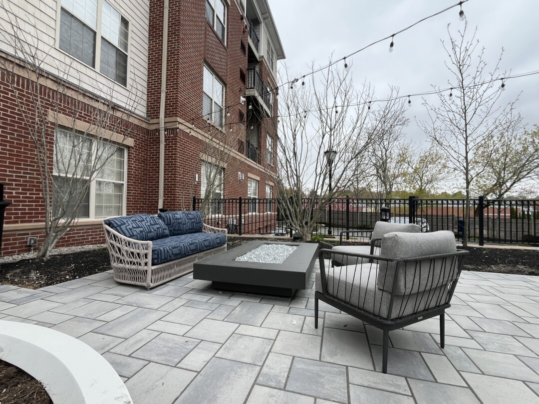 Outdoor lounge area with gray stone paver flooring, black metal and wicker seating with gray cushions, and a rectangular fire table; surrounded by landscaping, fencing, and brick building exterior.