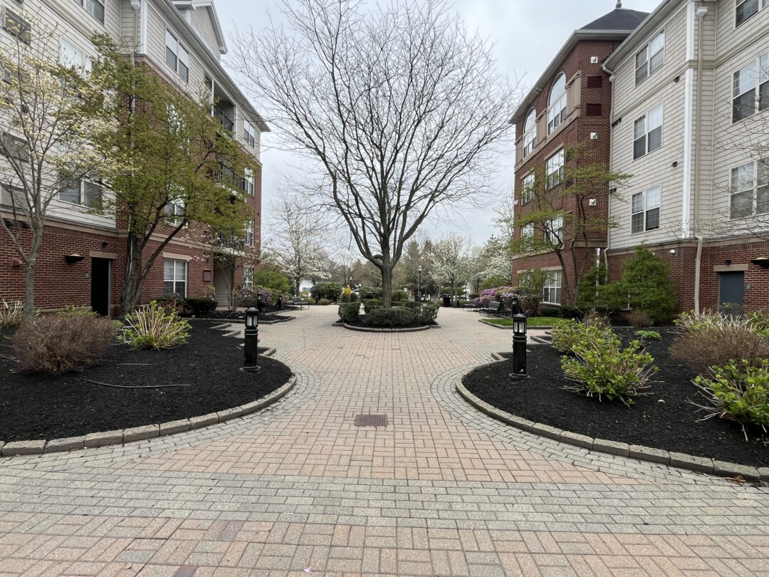 Landscaped courtyard with light brown and gray brick paver walkways, black pathway lighting, mulch beds with greenery, and surrounding red brick and beige-sided buildings.