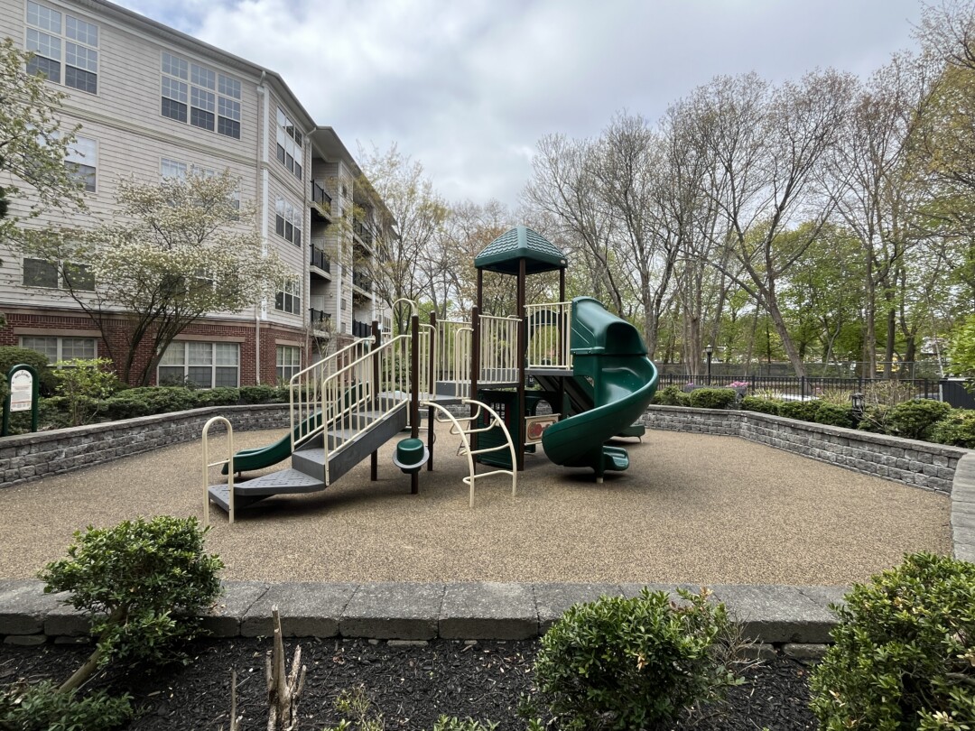 Outdoor playground with green and beige play structure on a tan surface; bordered by low stone walls, shrubs, and surrounded by residential buildings and trees.