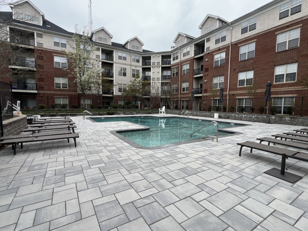 Outdoor pool with light blue water surrounded by gray stone pavers, lounge chairs, and umbrellas; enclosed by multi-story residential buildings with red brick and beige siding.