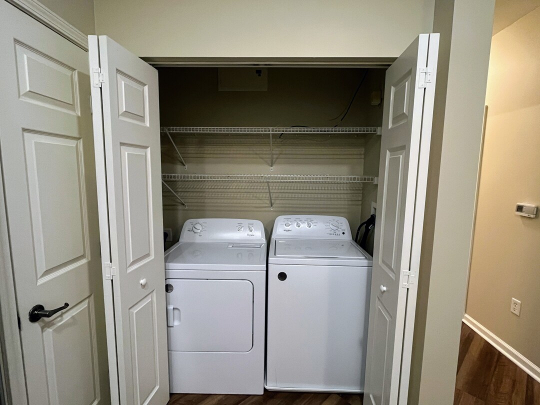 Laundry closet with white washer and dryer units, white walls, white ceiling, wood-style flooring, and white wire shelving above.