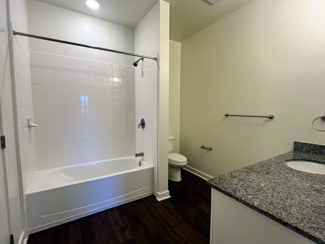 A bathroom with a white tub and shower surrounded by white tile, a toilet partially enclosed by a dividing wall, and a vanity with a countertop and sink, along with towel bars and dark wood flooring.