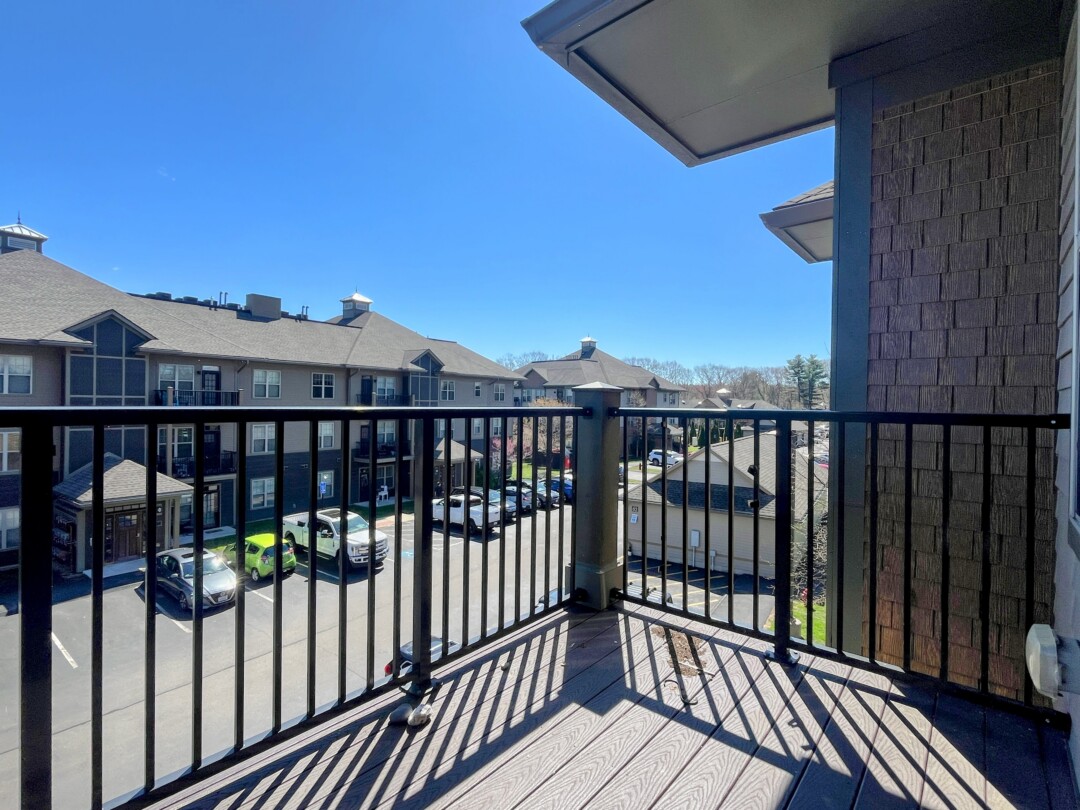 A balcony with wood flooring and a metal railing, overlooking a parking area and surrounding residential buildings under a clear blue sky.