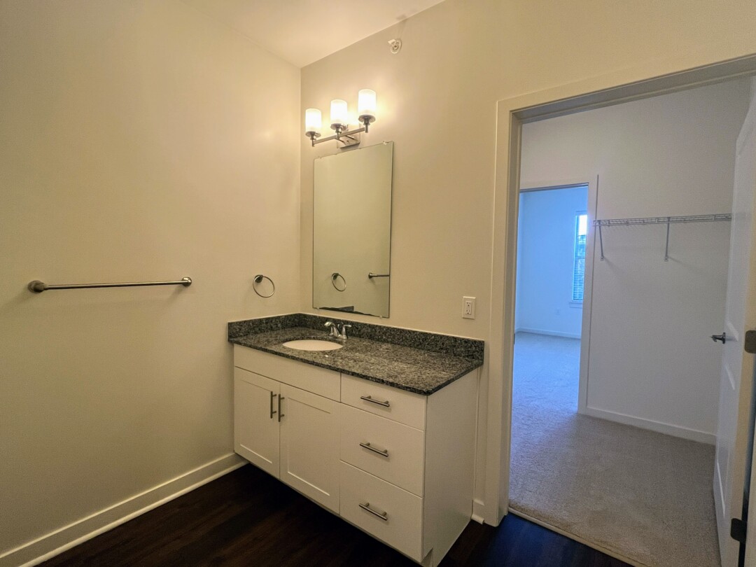 A bathroom with a vanity featuring white cabinets, a countertop with a sink, and a mirror with wall-mounted lighting above, along with towel bars and dark wood flooring, opening into a carpeted room with a window and an adjacent open area with a shelf and hanging rod.