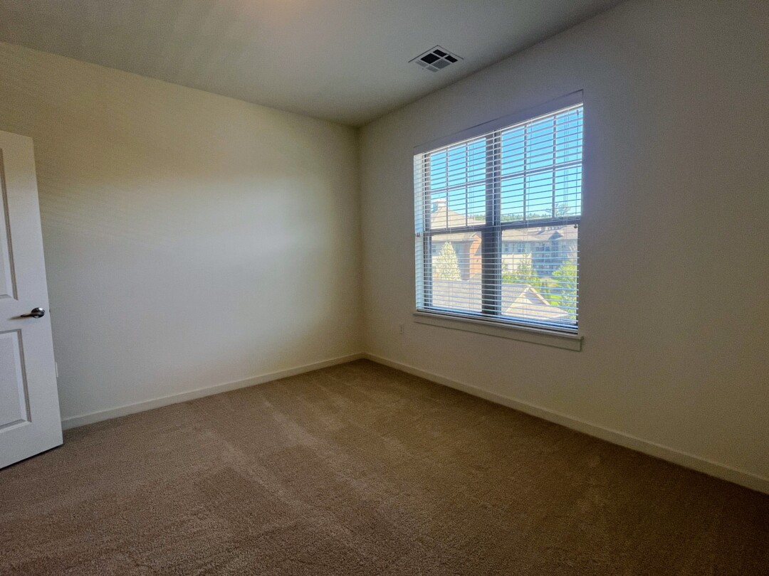 An empty bedroom with carpeted flooring and light-colored walls, featuring a window with blinds that lets in natural light and a door visible along one wall.