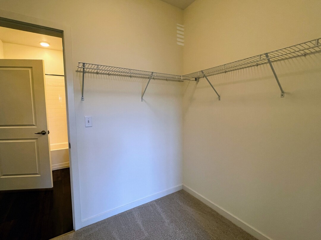 A bedroom closet with carpeted flooring, light-colored walls, and wire shelving along the walls for storage, with a doorway leading to a bathroom visible nearby.