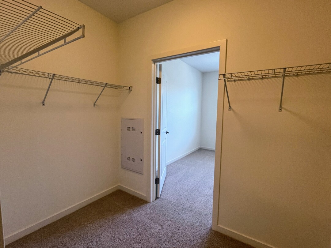 A carpeted bedroom closet with light-colored walls and wire shelving along the sides for storage, featuring a small wall panel and an open doorway leading to an adjacent room.”