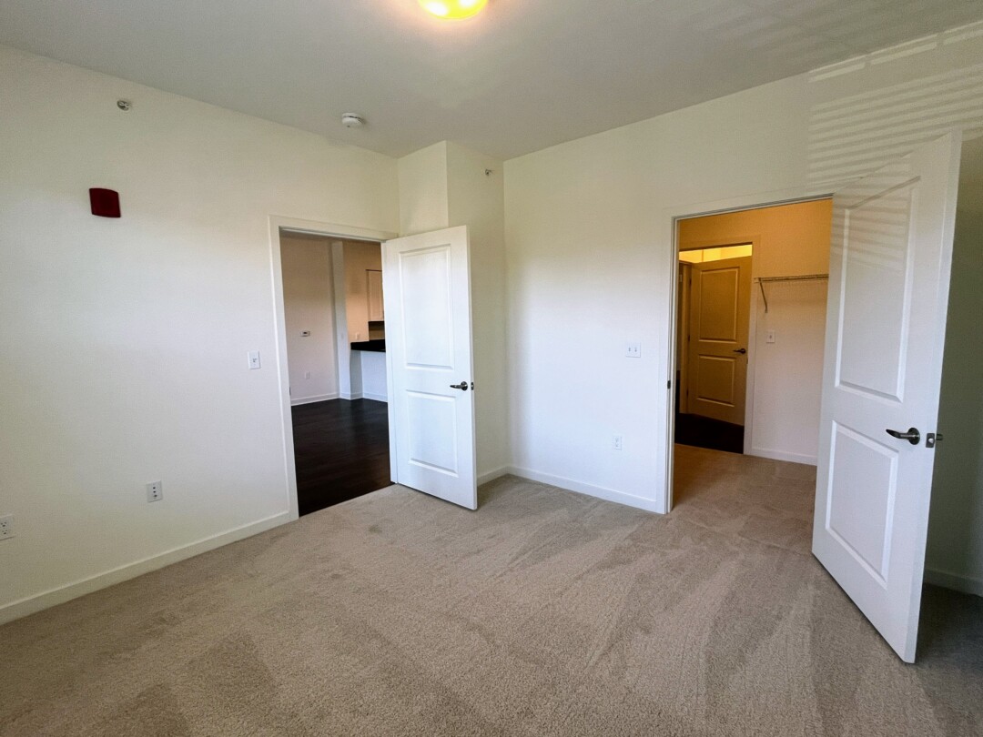 An empty bedroom with carpeted flooring and light-colored walls, featuring two open doorways—one leading to a living area with dark wood flooring and another to a closet or adjacent space—with ceiling-mounted lighting above.