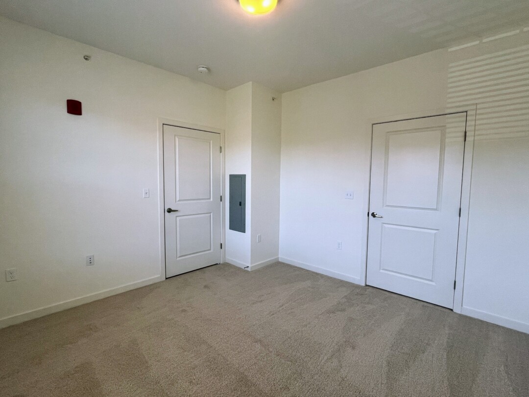 An empty bedroom with carpeted flooring and light-colored walls, featuring two closed doors and a small wall panel, with ceiling-mounted lighting above.