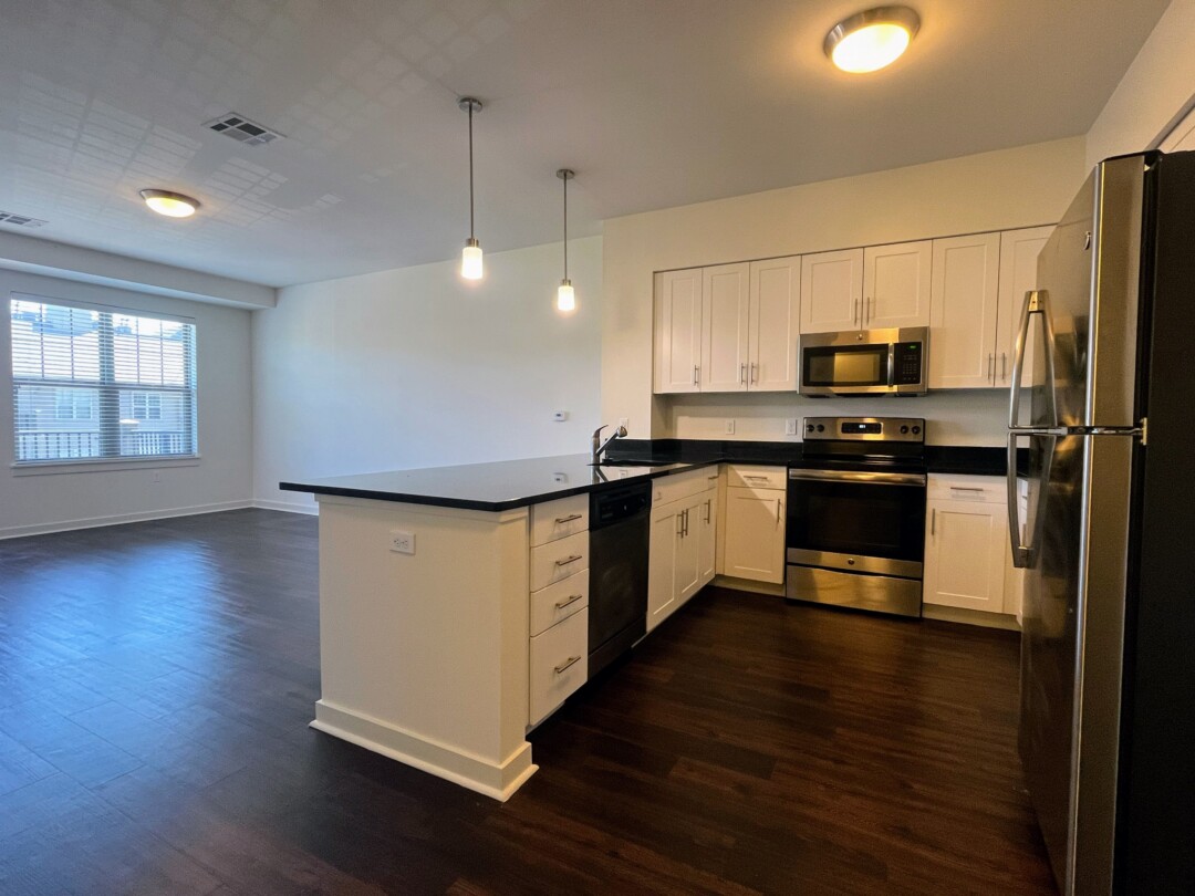 An open kitchen and living area with dark wood flooring, featuring white cabinets and dark countertops, stainless steel appliances including a refrigerator, oven, dishwasher, and over-the-range microwave, with two hanging light fixtures above the counter and a window in the adjacent living space.
